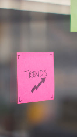 Vertical-Video-Close-Up-Of-Woman-Putting-Sticky-Note-With-Trends-Written-On-It-Onto-Transparent-Screen-In-Office